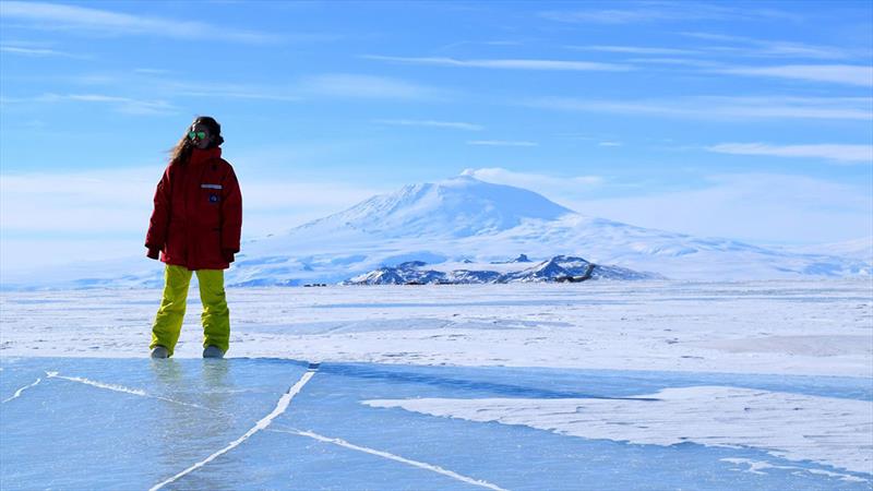 Catherine Walker stands on the McMurdo Ice Shelf in Antarctica in Oct 2014. In the background, U.S. Antarctic Program's McMurdo Station is visible at the base of Mt. Erebus, an active volcano, along with a C-130 aircraft delivering people & cargo - photo © Jacob Buffo/Georgia Tech/Dartmouth College